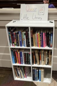 photo shows a small bookcase, filled with books, labelled queer little library at Flame Con 2025
