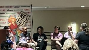 this photo of the flamecon panel described in the article shows a row of five people and a banner with a RESIST sign behind