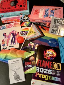 Emily's photo shows piled books, many stickers, and a progress pride flag as well as bookmarks and ephemera such as a mask, program and badge
